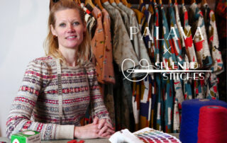 A smiling blonde woman dressed in brightly coloured fairisle jumper is sitting in front of a shop clothing rail with brightly coloured dresses behind her and a table with sewing items and a pantone book in front of her